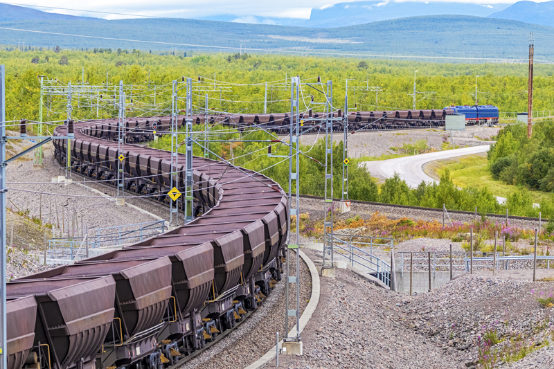 Loaded iron ore train snaking around curves through maze of electric poles and wires in Arctic Circle extreme terrain hauled by two electric locomotives